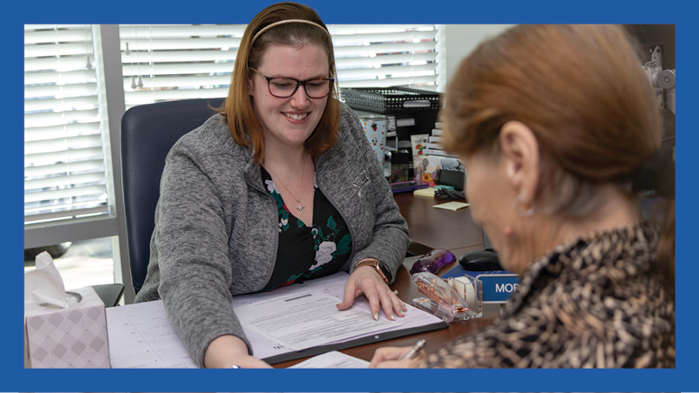 Person sitting at desk helping another person fill out paper